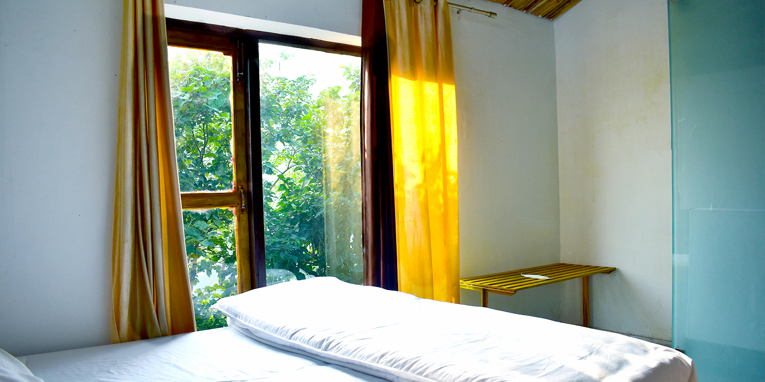 Zu sehen ist die Innenansicht eines Schlafzimmers. Im Vordergrund steht ein leeres Doppelbett. Dahinter ist ein großes Fenster sichtbar mit gelben Vorhängen, durch das die Sonne hereinscheint.